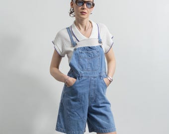 May include: Light blue denim overall shorts with a white short-sleeve top. The top has a white collar and purple trim. The model is wearing sunglasses and silver jewelry. The overall shorts have pockets and adjustable straps.