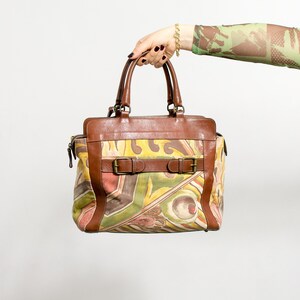 May include: A brown leather handbag with a colorful abstract pattern. The bag features two top handles, a rectangular shape, and a buckle closure. The bag has brown leather trim and a zipper closure.