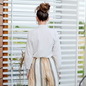 May include: A woman wearing a white button-down shirt and striped pants stands in front of a window with white blinds.