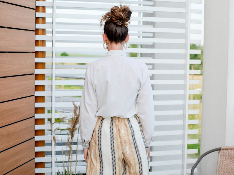 May include: A woman wearing a white button-down shirt and striped pants stands in front of a window with white blinds.