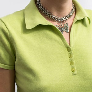K&ouml;nnte beinhalten: Ein limegr&uuml;nes Poloshirt mit einer silbernen Kettenkette mit einem Schmetterlingsanh&auml;nger. Das Shirt hat einen Knopfschlie&szlig;enverschluss.