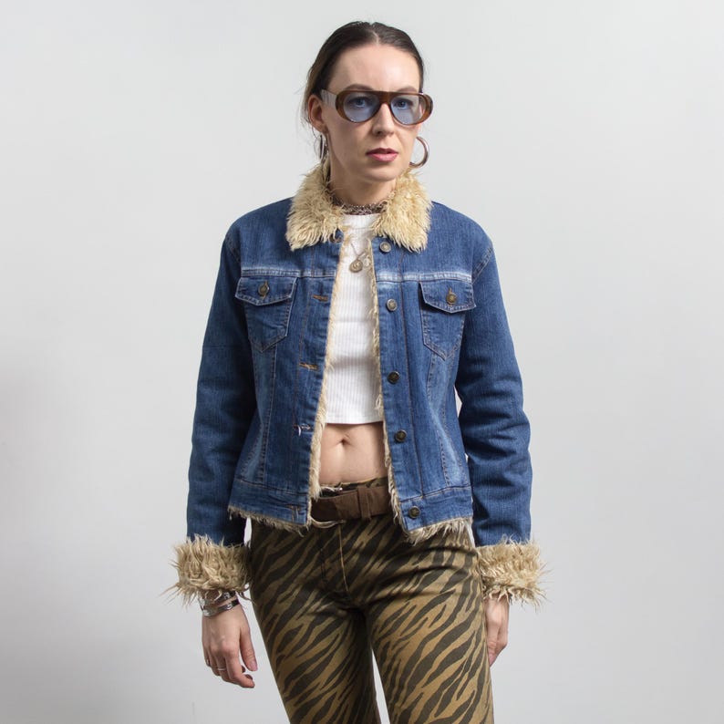 Peut inclure: Veste en jean bleue avec col et poignets en fausse fourrure beige. La veste a une fermeture boutonn&eacute;e et deux poches poitrine. La personne porte un pantalon z&eacute;br&eacute; marron et beige et des lunettes de soleil marron.