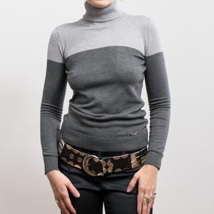 Könnte beinhalten: Ein grau-dunkelgrauer Rollkragenpullover. Der langärmlige Pullover hat einen gerippten Saum und ein kleines Logo unten. Die Person trägt einen schwarzen Gürtel mit silberner Schnalle und eine schwarze Hose.