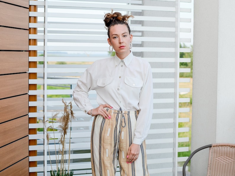 May include: A woman wearing a white button-down shirt with a pair of striped pants. She is standing in front of a window with white blinds.