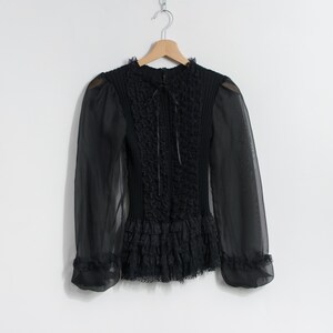Puede incluir: Blusa negra de manga larga con cuello alto y mangas transparentes. El corpi&ntilde;o presenta detalles texturizados intrincados y un dobladillo con volantes. La blusa est&aacute; colgada en una percha de madera sobre un fondo blanco.