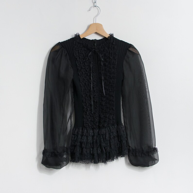 Puede incluir: Blusa negra de manga larga con cuello alto y mangas transparentes. El corpi&ntilde;o presenta detalles texturizados intrincados y un dobladillo con volantes. La blusa est&aacute; colgada en una percha de madera sobre un fondo blanco.