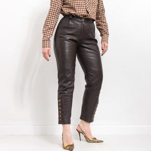 May include: Dark brown leather pants with gold button details down the sides. The pants have a high waist and a tailored fit. The model is wearing a brown and tan plaid button-down shirt and gold patterned heels.