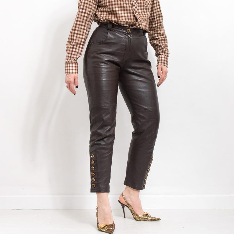 May include: Dark brown leather pants with gold button details down the sides. The pants have a high waist and a tailored fit. The model is wearing a brown and tan plaid button-down shirt and gold patterned heels.