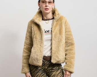 May include: A tan faux fur jacket with a high collar and a zipper closure. The jacket is worn over a white t-shirt with the word "ice" printed on it. The person is wearing brown zebra print pants and sunglasses.