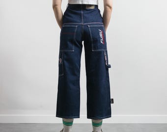 May include: Dark blue denim pants with white stitching and multiple pockets. The pants feature the brand name "FUBU" in red and white on the back. The pants have a wide leg design and are worn with sneakers and striped socks.