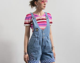 May include: Light blue denim overall shorts with a patch and star-patterned cuffs. The shorts are paired with a striped pink, red, and white short-sleeved top. The person is wearing pink sunglasses and silver jewelry.