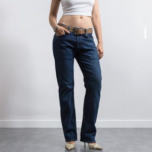 May include: A pair of dark blue denim jeans featuring a straight-leg cut, a classic five-pocket design, and a button fly closure. The jeans are worn with a brown leather belt that has a large, rectangular metal buckle with a decorative cutout.