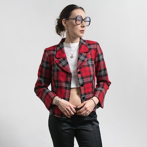 Könnte beinhalten: Ein rot-schwarz karierter Blazer mit klassischem Kragen und zwei schwarzen Knöpfen. Die kurze Jacke wird mit einer schwarzen Hose kombiniert. Das Muster des Blazers ist ein traditionelles Tartanmuster.