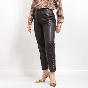 May include: Dark brown leather pants with gold button details on the lower leg. The pants are paired with gold-colored pointed-toe heels. The person is wearing a brown and tan plaid button-down shirt.