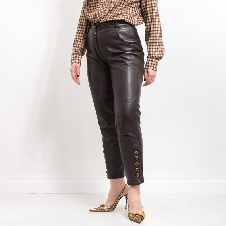 May include: Dark brown leather pants with gold button details on the lower leg. The pants are paired with gold-colored pointed-toe heels. The person is wearing a brown and tan plaid button-down shirt.