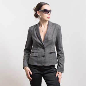 Puede incluir: Una blazer gris a cuadros con ribete negro y cierre de un botón. La blazer tiene solapas con muescas y un pequeño bolsillo en el lado izquierdo. La modelo lleva pantalones negros y gafas de sol.