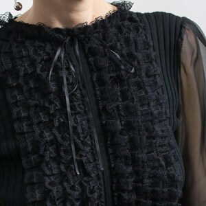 Puede incluir: Top negro con frente de encaje texturizado y mangas transparentes. El top presenta un cuello con volantes y un lazo de cinta de sat&eacute;n. La tela tiene una textura acanalada en los laterales y la espalda.