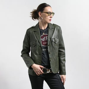 Può includere: Giacca in pelle verde oliva con quattro tasche frontali e bottoni. La giacca è abbinata a una maglietta Hard Rock Cafe, pantaloni neri e una cintura fantasia. La persona indossa occhiali da sole e orecchini a cerchio argentati.