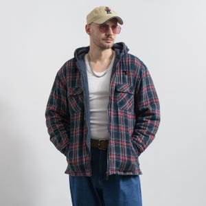 Könnte beinhalten: Eine beige Baseballkappe mit kleinem Motiv, rosagetönte Sonnenbrille, ein weißes Unterhemd und eine blau-rot karierte Kapuzenjacke mit Steppfutter. Die Jacke hat einen Frontreißverschluss, zwei Brusttaschen mit Klappen und eine Kapuze mit Kordelzug.
