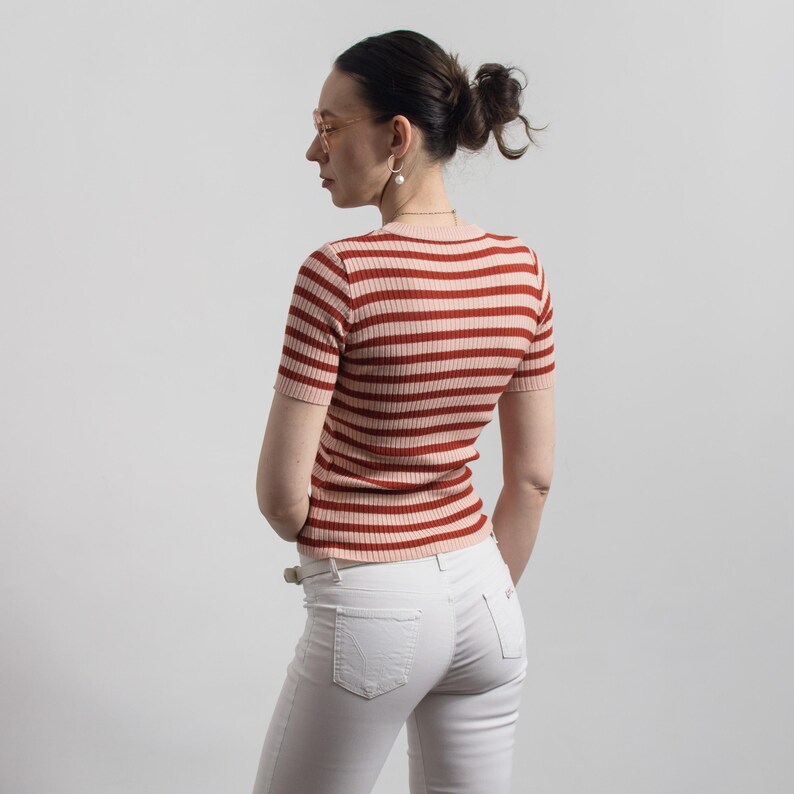 May include: A short-sleeved, ribbed knit top with horizontal stripes in shades of red and light pink. The top is paired with white jeans. The model is wearing glasses and earrings.