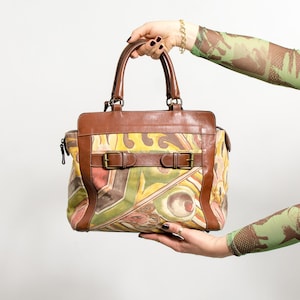 May include: A brown leather handbag with a colorful abstract design. The bag features a top handle, a shoulder strap, and two buckle closures. The bag's design includes yellow, green, and red colors.