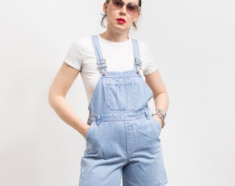 Puede incluir: Un short vaquero de mezclilla azul claro con tirantes ajustables. Los tirantes tienen hebillas plateadas. La modelo lleva una camiseta blanca y gafas de sol.
