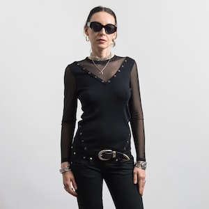 Puede incluir: Una blusa negra de manga larga con mangas de malla transparente y un escote en V acentuado con ojales de metal plateado. La prenda incluye detalles de ojales adicionales a lo largo de los paneles laterales y se combina con pantalones negros.