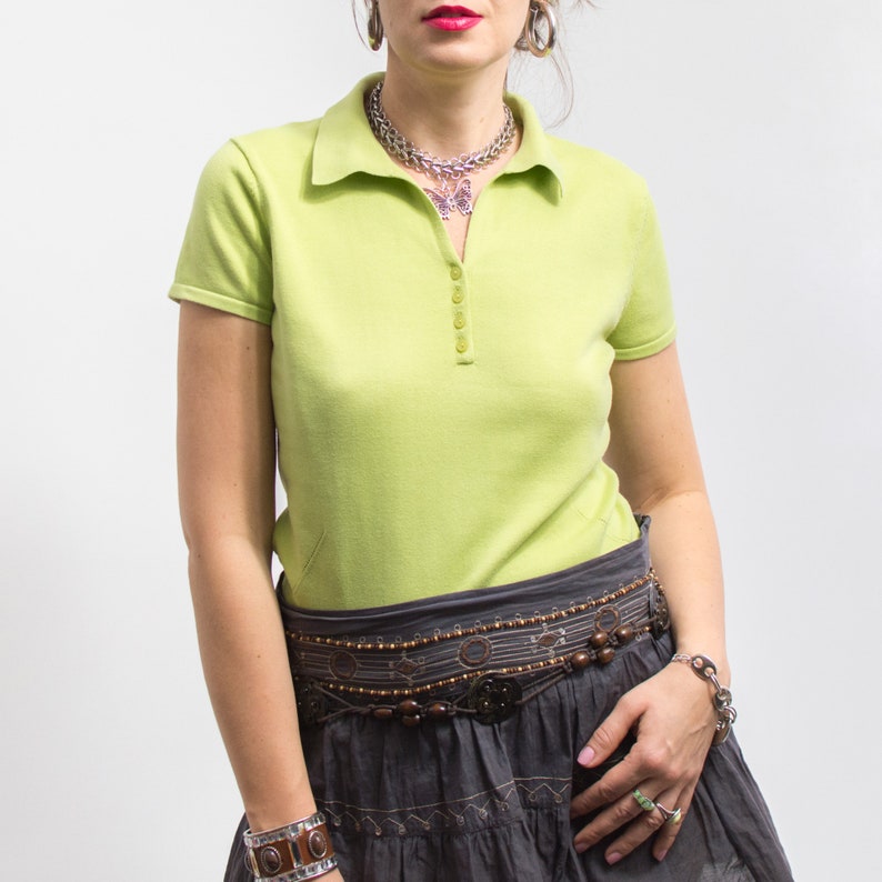 K&ouml;nnte beinhalten: Ein kurz&auml;rmliges Poloshirt in Limettengr&uuml;n mit Knopfleiste und einer silbernen Schmetterlingskette. Das Shirt wird mit einem dunkelgrauen Rock mit einem braunen Perleng&uuml;rtel kombiniert.