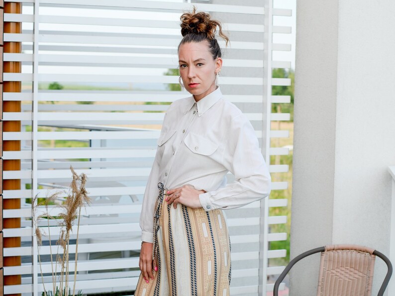 May include: A woman wearing a white button-down shirt with a long sleeve and a brown and white striped skirt. She is standing in front of a white slatted wall.
