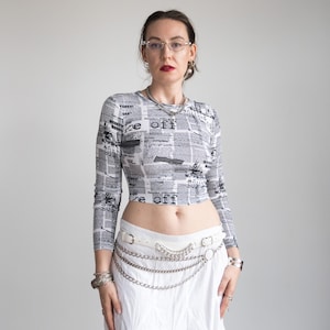 Op de afbeelding: Een cropped shirt met lange mouwen met een krantenprint in grijstinten en wit. Het shirt heeft de tekst "re off" op verschillende plaatsen. Het model draagt een witte riem met zilveren kettingaccenten en een witte rok.