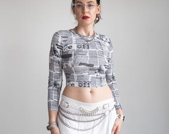 Op de afbeelding: Een cropped shirt met lange mouwen met een krantenprint in grijstinten en wit. Het shirt heeft de tekst "re off" op verschillende plaatsen. Het model draagt een witte riem met zilveren kettingaccenten en een witte rok.