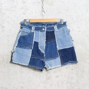Op de afbeelding: Denim shorts met een patchwork ontwerp in verschillende blauwtinten. De shorts hebben een gerafelde zoom en hangen aan een houten hanger. De shorts hebben een knoopsluiting en zakken.