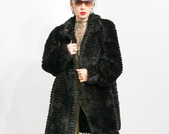 May include: A black faux fur coat with a textured, patterned design. The coat has a classic collar and a knee-length cut. The model is wearing sunglasses and jewelry. The coat is a stylish outerwear piece.