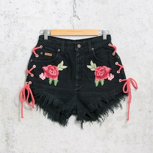Może przedstawiać: Czarne szorty dżinsowe z postrzępionymi krawędziami, zdobione haftowanymi czerwonymi różami i zielonymi liśćmi. Boki mają różowe sznurowanie. Szorty wiszą na drewnianym wieszaku.