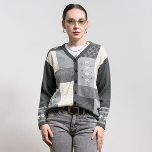 May include: A gray, white, and black patchwork cardigan sweater with a button-up front. The sweater features floral embroidery and a checkerboard pattern on the upper right panel. The model is wearing gray jeans and a black belt.