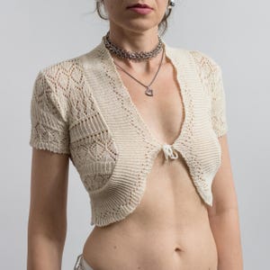 May include: A cream-colored, short-sleeved knit bolero cardigan featuring an intricate openwork lace pattern, ribbed trim along the edges, and a single tie closure at the center front. The garment is cropped, ending above the waistline.