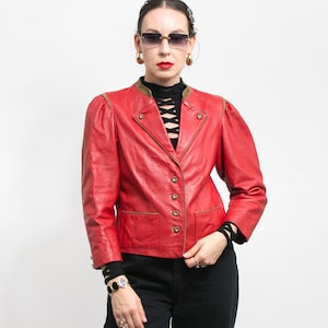 May include: A red leather jacket with gold-colored buttons and trim. The jacket has a classic design with a notched collar, two front pockets, and padded shoulders. The model is wearing black pants and a black top.