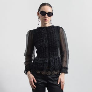 Puede incluir: Blusa negra con mangas largas transparentes, corpi&ntilde;o con volantes y detalle decorativo en la cintura. La blusa tiene un cuello alto y una silueta entallada. La modelo lleva gafas de sol negras y pendientes. La blusa se combina con pantalones negros.