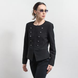 Op de afbeelding: Zwarte jas in militaire stijl met zilverkleurige knopen en decoratieve stiksels. De jas heeft een getailleerd silhouet en een hoge kraag. De jas wordt gedragen met een zwarte broek.