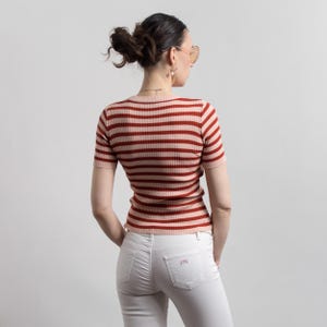 May include: A short-sleeved, striped knit top in shades of red and pink. The top is paired with white jeans. The model is wearing sunglasses and has their hair up in a bun. The top has a round neckline.