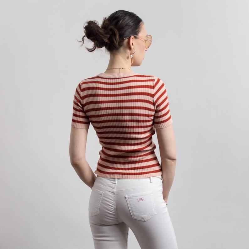 May include: A short-sleeved, striped knit top in shades of red and pink. The top is paired with white jeans. The model is wearing sunglasses and has their hair up in a bun. The top has a round neckline.