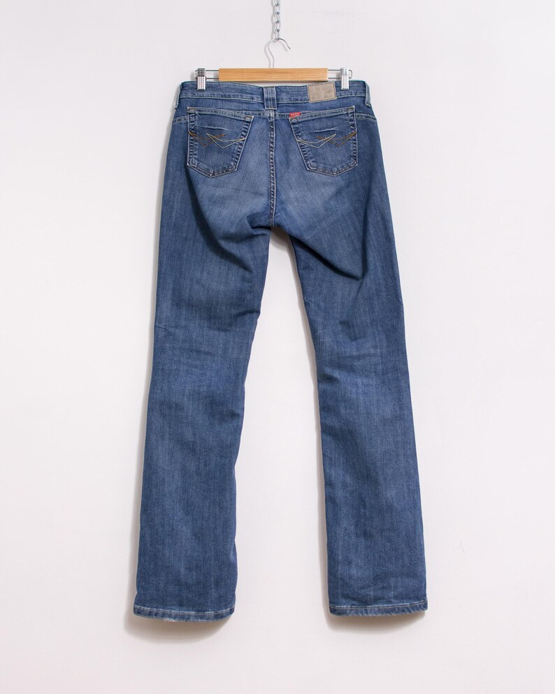 Peut inclure: Jean bleu en denim suspendu &agrave; un cintre en bois. Le jean a une conception classique &agrave; cinq poches, avec des coutures d&eacute;coratives sur les poches arri&egrave;re. Une &eacute;tiquette rouge est visible sur la ceinture arri&egrave;re. Le jean est de couleur moyenne.