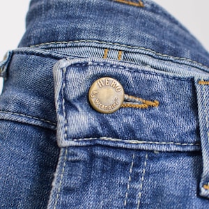 Peut inclure: Gros plan sur un jean en denim bleu avec une fermeture boutonn&eacute;e. Le bouton dor&eacute; porte l'inscription "HERO" et "by Wrangler". Le jean pr&eacute;sente des coutures visibles et un design classique.
