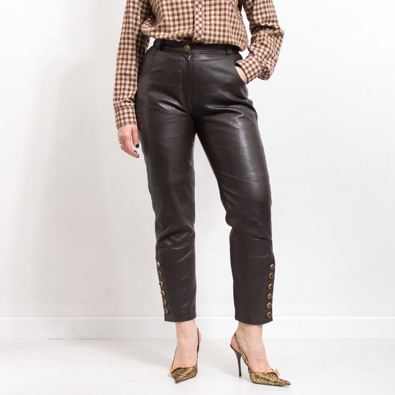 May include: Dark brown leather pants with gold button details along the ankles. The pants are high-waisted and have a classic, tailored fit. The model is wearing a brown plaid button-down shirt and gold heels.