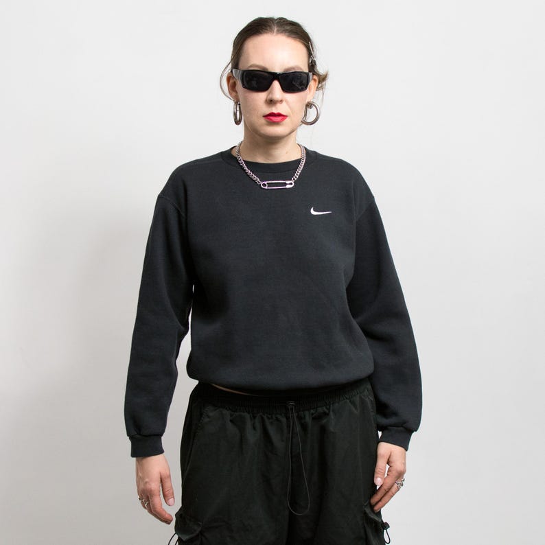 May include: A black sweatshirt with a small white Nike logo on the chest. The person is wearing black sunglasses, hoop earrings, and a silver chain necklace. The person is also wearing black cargo pants.