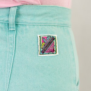 Puede incluir: Un par de pantalones vaqueros de color azul claro con un peque&ntilde;o parche rectangular en el bolsillo trasero derecho. El parche es blanco con detalles en rosa, amarillo y azul y dice "Petrolicious" en rosa.