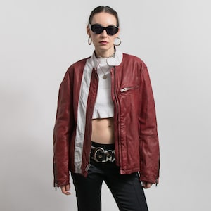 May include: A burgundy leather jacket with a white stripe down the front. The jacket has a zip closure, side pockets, and a white collar. The model is wearing black sunglasses and black trousers.