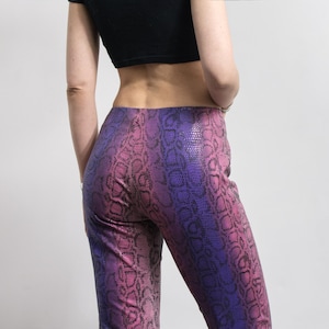 May include: High-waisted pants with a snakeskin pattern in shades of pink and purple. The pants have a fitted silhouette and a subtle sheen. The fabric appears to be a stretchy material.