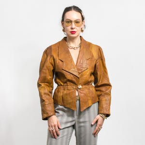 Op de afbeelding: Een vintage bruine leren jas met een uniek patchwork ontwerp. De jas heeft een reverskraag, een getailleerde taille en een enkele knoopsluiting. Het model draagt een grijs-witte gestreepte broek en gouden sieraden.