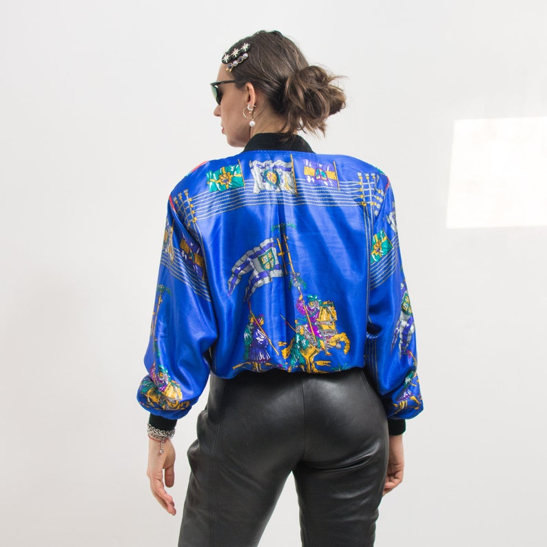May include: A blue satin bomber jacket with a colorful print of knights on horseback. The jacket has a zipper closure and a stand-up collar.